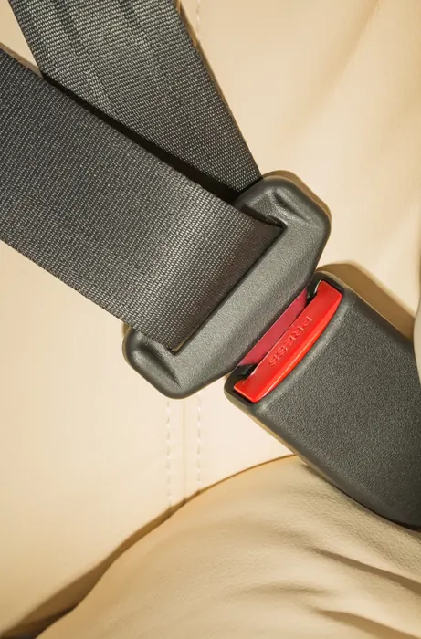 Seat belt webbing entering a black buckle with a red release button, photographed against a light vehicle seat.